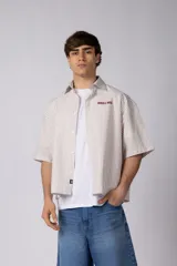 Camisa de manga corta a rayas beige y blancas, de corte boxy y liviana, con cuello camisero, cierre frontal de botones y pequeño texto bordado "BUILD IT BOLD" en el pecho.