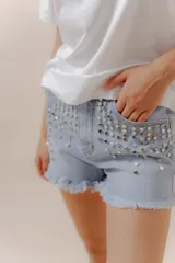 Short de jean celeste con tachas metálicas plateadas, tiro medio, terminaciones desflecadas, cierre frontal y bolsillos funcionales.