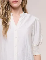 Blusa blanca de crepé con cuello mao y escote en V, cierre frontal con botones, mangas cortas abullonadas con puños elastizados en punto smock y calce semi holgado.