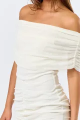 Vestido corto de mesh color blanco, con diseño off-shoulder y escote con volados. Presenta un ajuste drapeado y fruncido en la parte central de la falda, finalizando con un ruedo con volado sutil.