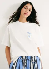 Remera blanca de manga corta, corte oversized, cuello redondo y pequeño estampado azul en el pecho con diseño de palmera y texto.