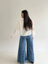 Blusa de brodery color blanco, con cuello redondo, mangas largas abullonadas con puños elásticos y cierre frontal con botones forrados. Presenta detalles de puntillas verticales en el frente.