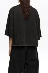 Remera corta de corte oversize color negro, confeccionada en mezcla de algodón. Presenta cuello redondo, mangas cortas amplias y un gran bordado frontal con diseño estilo etiqueta y texto.
