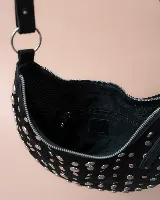 Bolso de mano tipo hobo, confeccionado en gamuza color marrón oscuro, decorado con tachas metálicas circulares en toda su superficie. Cuenta con un asa corta integrada y un diseño curvo distintivo.