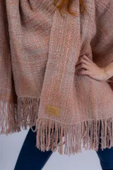 Ruana artesanal tejida en telar manual, con textura rústica y terminación en flecos. Presenta un diseño liviano en tonos rosados y naranjas, con etiqueta de marca en el frente.