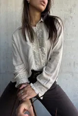 Camisa de color beige con diseño romántico, presenta una pechera central con encaje floral y botones frontales. Posee mangas largas con puños decorados con volados y encaje a juego.