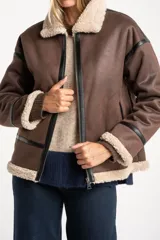 Campera tipo gamulán de corte oversize, color marrón con interior y cuello de corderito sintético en tono crema. Presenta detalles de ribetes en cuero sintético negro a lo largo de la prenda, cierre frontal metálico y puños tejidos.