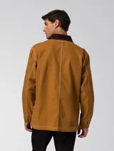 Chaqueta tipo chore de lona color marrón, con cuello camisero en contraste color marrón oscuro, cierre frontal con botones, cuatro bolsillos de parche frontales y costuras a la vista en color blanco.