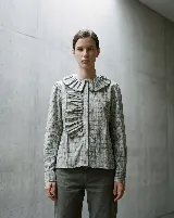 Camisa de algodón con estampado a cuadros en tonos grisáceos. Presenta un cuello amplio con volados, detalles fruncidos en el frente, cierre frontal con botones y mangas largas con puños abotonados.