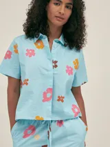 Camisa de manga corta confeccionada en tejido seersucker celeste con estampa floral en tonos naranja, rosado y marrón. Posee cuello camisero, cierre frontal con botones anacarados y un corte recto y holgado.