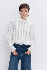 Camisa de algodón color blanco con detalles de encaje broderie y alforzas verticales. Presenta cuello alto con volados, cierre frontal de botones y mangas largas con puños acampanados.