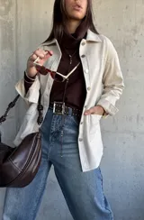 Chaqueta tipo sobrecamisa color beige, con cierre frontal de botones metálicos, cuello clásico y bolsillos frontales. Presenta un diseño de corte recto y mangas largas con puños abotonados.