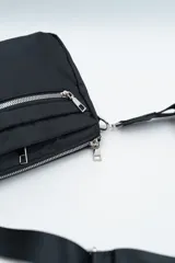 Bandolera de diseño compacto en color negro, confeccionada en material sintético resistente. Cuenta con un compartimento principal con cierre y un bolsillo frontal con cierre metálico visible. Incluye una correa larga ajustable y extraíble para llevar al hombro o cruzada.