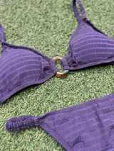 Conjunto de bikini color violeta confeccionado en tela texturada. El corpiño es de estilo top con una hebilla ovalada marrón en el centro y breteles finos. La bombacha es de corte colaless con detalle de frunces en los laterales.