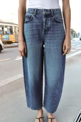 Jeans de tiro alto con diseño barrel, confeccionados en denim azul. Presentan cinco bolsillos, cierre frontal con botones y detalle de costuras verticales en el frente y la espalda. Su corte es holgado en la pernera y se estrecha hacia el tobillo.