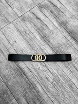 Cinto negro de cuero sintético con hebilla dorada de tres aros entrelazados.