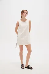 Vestido corto de algodón color blanco, de corte holgado, sin mangas y con cuello a la base. Presenta detalles de cordones cruzados y ojalillos en los laterales.