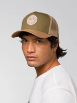 Gorro trucker de color verde militar, con visera curvada marrón claro, panel frontal rígido y red trasera. Cuenta con un parche circular bordado en la parte delantera y cierre ajustable snapback.