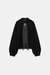 Chaqueta de punto negra con cuello redondo, cierre frontal con cremallera metálica y detalle de lazada en tejido a cuadros en contraste. Presenta mangas largas abullonadas con puños elásticos y bajo elástico.