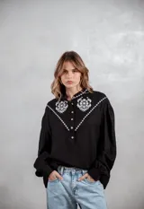Camisa negra de manga larga con bordados blancos en la parte superior delantera y hombros. Presenta cuello mao, cierre frontal con botones y mangas con puños ajustados.