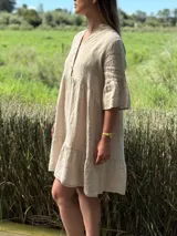 Vestido corto de lino color beige claro, con cuello mao, escote con botones de nácar, mangas tres cuartos con volados y ruedo con volado.