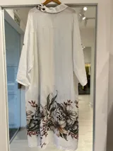 Vestido largo estilo camisero color blanco, con cierre frontal de botones, bolsillos superiores y un gran estampado floral en tonos marrones y verdes en la parte inferior y en el bolsillo.