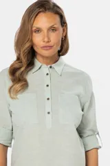 Camisa blanca de manga larga con cuello y tapeta de medio botón. Presenta dos bolsillos frontales con tapa y botones metálicos. Las mangas tienen presillas para sujetar el puño enrollado.