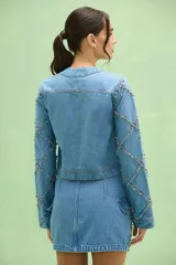 Chaqueta denim de mujer, confeccionada en jean azul claro, con cuello redondo y cierre frontal. Destaca por apliques bordados florales con pedrería y flecos de cristales en la parte frontal, además de detalles de brillos y costuras decorativas a lo largo de las mangas.