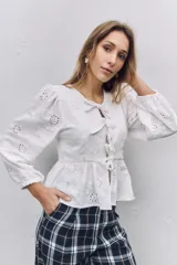 Blusa blanca de tela broderie con diseño de flores caladas, mangas largas abullonadas con puños elásticos y cierre frontal mediante lazos decorativos.