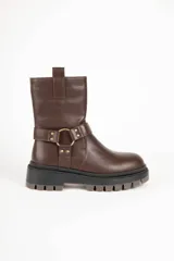 Bota estilo biker de cuero sintético color marrón, con suela tipo plataforma dentada negra. Presenta una tira decorativa en el tobillo con una argolla metálica dorada y apliques de tachas.