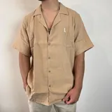 Camisa de manga corta confeccionada en muselina de algodón, con cuello tipo cubano y corte oversize. Presenta cierre frontal con botones y un pequeño bordado en el pecho.