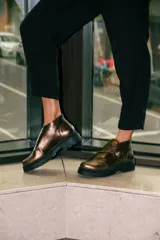 Botines de cuero metalizado en tono oscuro con acabado brillante. Presentan un diseño de corte bajo con una hebilla lateral decorativa y suela de goma negra con plataforma dentada.