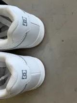 Championes DC modelo Manteca, color blanco con detalles en gris, cordones anchos y suela de goma marrón. Presentan el logo de DC en la lengüeta y los laterales.