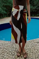 Falda midi de corte cruzado con diseño tipo pareo, presenta un nudo decorativo en la parte delantera y cierre de cremallera oculta en la espalda. El tejido cuenta con un estampado geométrico abstracto en tonos bordó, negro, amarillo y crema.