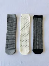 Pack de tres pares de medias de caña media. Un par gris con rayas horizontales negras, un par blanco con lunares negros y un par negro con diseño de espiga en gris.