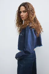 Campera cropped de denim de algodón rígido, color azul, con cuello, cierre frontal de botones y hombros caídos. Cuenta con bolsillos superiores con solapa y botón, puños abotonados y dobladillo deshilachado.