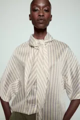 Camisa de corte holgado confeccionada en mezcla de lyocell, con estampado de rayas verticales en tonos neutros y azules. Presenta cuello redondo con lazada frontal, cierre con botones ocultos y mangas amplias por debajo del codo.
