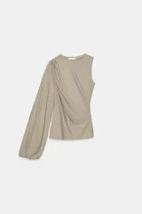 Top asimétrico color beige, con cuello redondo, una manga sisa y una manga larga abullonada. Presenta detalles de pliegues en el frente.