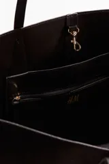 Bolso tipo shopper de color marrón oscuro o negro, con forma curva y dos asas de mano largas. Presenta costura central vertical y detalles de costura en forma de diamante donde se unen las asas al cuerpo del bolso.