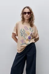 Remera color tostado de corte holgado, con cuello redondo y mangas cortas. Presenta un estampado gráfico frontal de estilo veraniego con ilustraciones de una langosta, conchas, limón, una copa y la inscripción 'Summer Club'.