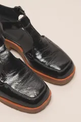 Zapato tipo guillermina de cuero charol color marrón oscuro, con tira T y hebilla ajustable en el lateral. Posee puntera cuadrada y suela de goma color caramelo con taco bajo.