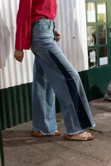 Pantalón de denim tiro alto con corte wide leg, lavado medio con desgastado suave, costuras clásicas y terminación en ruedo deshilachado. Diseño de panel lateral en tono más oscuro.