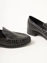 Mocasines de cuero negro con costuras pespunteadas en hilo blanco a contraste, diseño fruncido en la puntera, talón plano con pequeño tacón y apliques sutiles en charol.