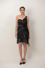 Vestido corto negro confeccionado en gasa drapeada sobre seda natural, con diseño asimétrico de un solo hombro. Presenta detalles de bordado artesanal con lentejuelas y piedras, y terminación de encaje en el ruedo de la falda.