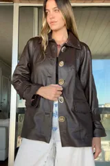 Tapado de cuero marrón con diseño artesanal, cuello camisero con detalles de pasamanería en gamuza, cierre frontal con botones de guampa y bolsillos delanteros con bordes decorativos. Presenta un corte cómodo y versátil.