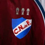 Camiseta de arquero Umbro del Club Nacional de Football, color bordó con detalles en blanco y azul. Presenta cuello en V, sponsors estampados y escudo del club en el pecho.