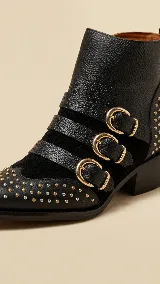 Botas cortas de cuero negro con diseño estilo western, decoradas con múltiples tachas metálicas en tono dorado y plateado. Presentan tres correas horizontales con hebillas doradas sobre el empeine y cierre lateral.