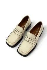 Mocasín color marfil de cuero, con detalle de tachas plateadas en la parte delantera y suela de goma negra.