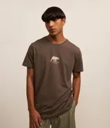 Remera marrón de corte clásico, confeccionada en algodón, con cuello redondo y manga corta. Presenta un estampado pequeño de un oso caminando sobre un paisaje.