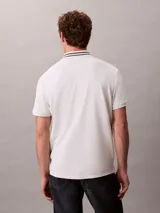 Chomba de piqué blanca Calvin Klein, de corte regular y manga corta, con cuello clásico con líneas grises en contraste y cartera de dos botones. Cuenta con el monograma CK bordado discretamente en el pecho.